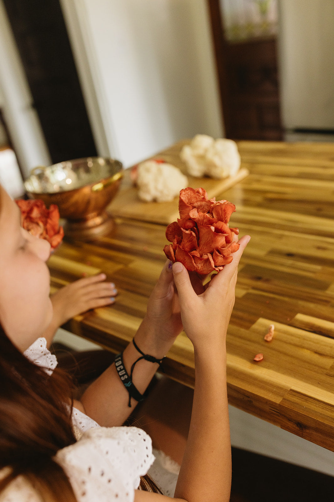 A hand holds a cluster of vibrant coral-pink oyster mushrooms above a striped bamboo cutting board in a sunlit kitchen, with white lion's mane mushrooms and a golden brass bowl visible on the wooden surface below.