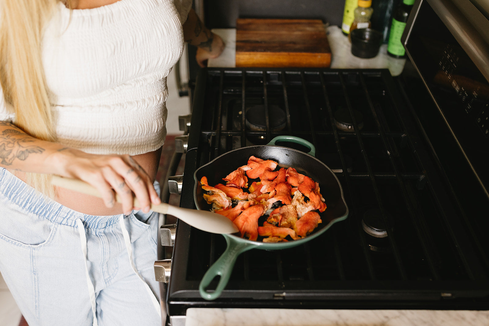 A blonde woman in a white crop top and light blue jeans stands at a black gas stove, cooking vibrant orange bell peppers and sliced mushrooms in a dark skillet with a mint green handle, her tattooed forearm visible as she tends to the sizzling vegetables in a sunlit kitchen with wooden cutting board and bottles visible on the counter.