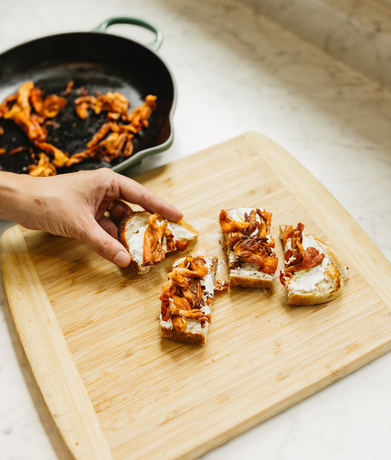 A hand reaches for golden-brown sautéed oyster mushroom crostini on a bamboo cutting board, with crispy caramelized mushroom strips spread over cream cheese on toasted bread slices, and a cast iron skillet with green handles containing more cooked mushrooms in the background.