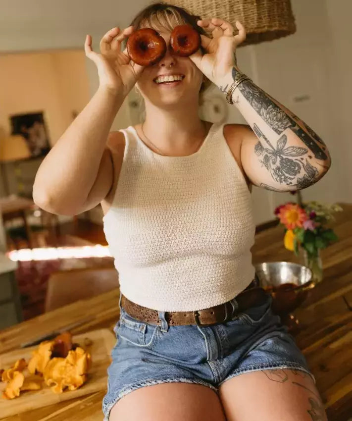 A smiling woman with blonde hair and floral arm tattoos playfully holds two rings of golden-yellow chanterelle mushrooms up to her eyes like binoculars in a sunlit kitchen, wearing a white knit crop top and denim shorts, with fresh mushrooms scattered on a wooden cutting board and colorful flowers visible in the background.