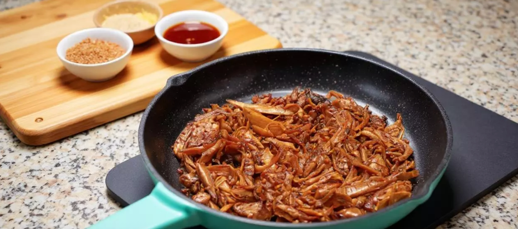 Shredded oyster mushrooms sautéed to golden-brown perfection in a black cast iron skillet with turquoise handle, garnished with sesame seeds, served on granite countertop with wooden cutting board holding white bowls of brown seasoning and dark sauce in the background.