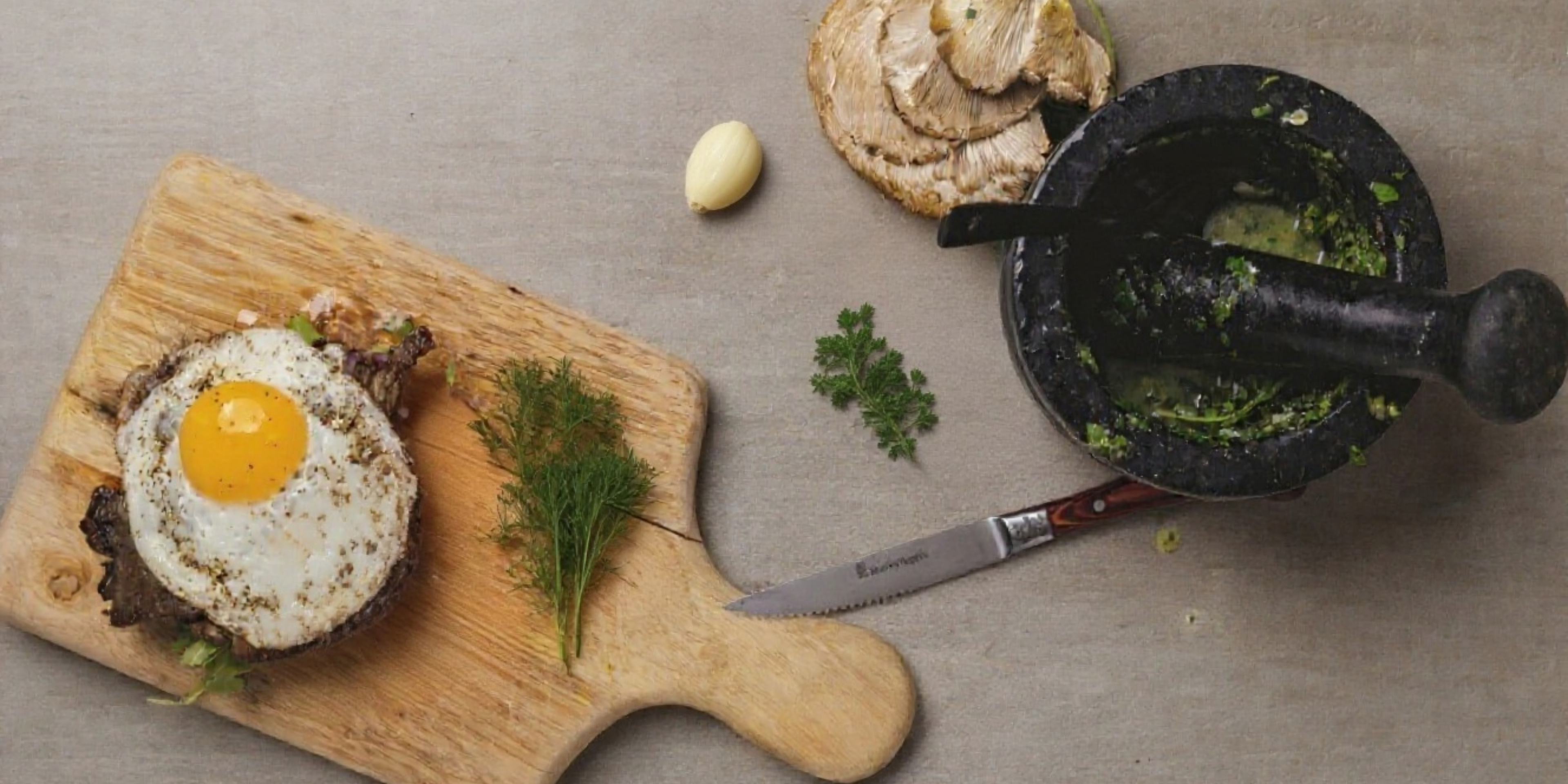 A sunny-side-up egg tops a crispy mushroom toast on a wooden cutting board, garnished with fresh dill, alongside a black marble mortar and pestle filled with green herb paste, a serrated knife, garlic clove, and whole portobello mushroom on a gray surface.